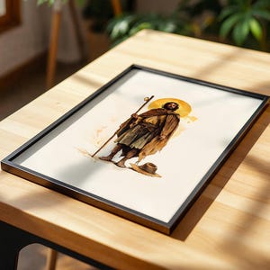 May include: A framed art print featuring a watercolour-style illustration of a bearded figure in a brown cloak and tunic, holding a wooden staff. The figure has a golden halo behind their head and stands near a small hat on a light background.