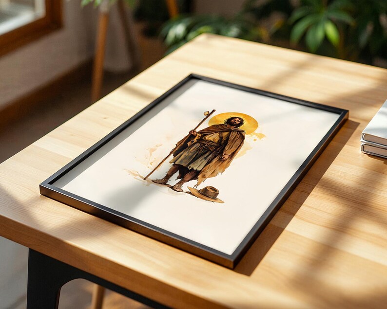 May include: A framed art print featuring a watercolour-style illustration of a bearded figure in a brown cloak and tunic, holding a wooden staff. The figure has a golden halo behind their head and stands near a small hat on a light background.