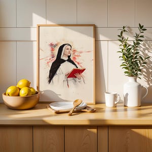 K&ouml;nnte beinhalten: Ein gerahmter Kunstdruck mit einer Aquarell-Illustration einer Nonne in schwarz-wei&szlig;em Habit, die ein rotes Buch h&auml;lt, vor einem leuchtend roten und orangefarbenen Sonnenstrahlen-Hintergrund, pr&auml;sentiert in einem hellen Holzrahmen auf einer K&uuml;chentheke.