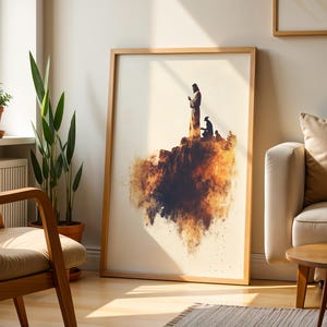 May include: A framed wall art print featuring a silhouette of a standing figure and a kneeling figure on a rocky cliff edge, rendered in warm orange, brown, and black watercolour-style splashes against a cream-coloured background.