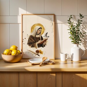 May include: A framed wall art print featuring a watercolour illustration of a figure in a brown habit and head covering writing in a book with a golden halo, a dove, and a quill pen, displayed on a wooden counter with lemons and a vase.