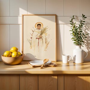 K&ouml;nnte beinhalten: Ein gerahmter vertikaler Druck mit einer Aquarellillustration einer Figur in wei&szlig;en Gew&auml;ndern mit einem goldenen Heiligenschein, die einen Palmwedel h&auml;lt. Der Druck steht auf einer Holztheke neben einer Schale mit gelben Zitronen, einem wei&szlig;en Teller, einem Holzl&ouml;ffel und einem wei&szlig;en Krug.