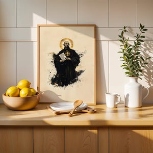 May include: A framed wall art print featuring a black and white illustration of a monk with a golden halo, holding a document with a cross emblem. The print is displayed on a wooden counter next to a bowl of yellow lemons and a white vase.