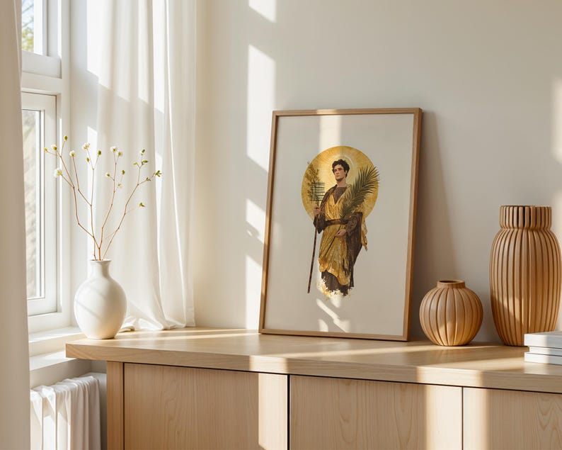 Op de afbeelding: Een ingelijste kunstdruk met een figuur in gouden gewaden die een palmtak en een staf vasthoudt, tegen een cirkelvormige gouden halo. De print staat op een houten oppervlak naast een witte vaas met dunne takken en twee geribbelde houten vazen.
