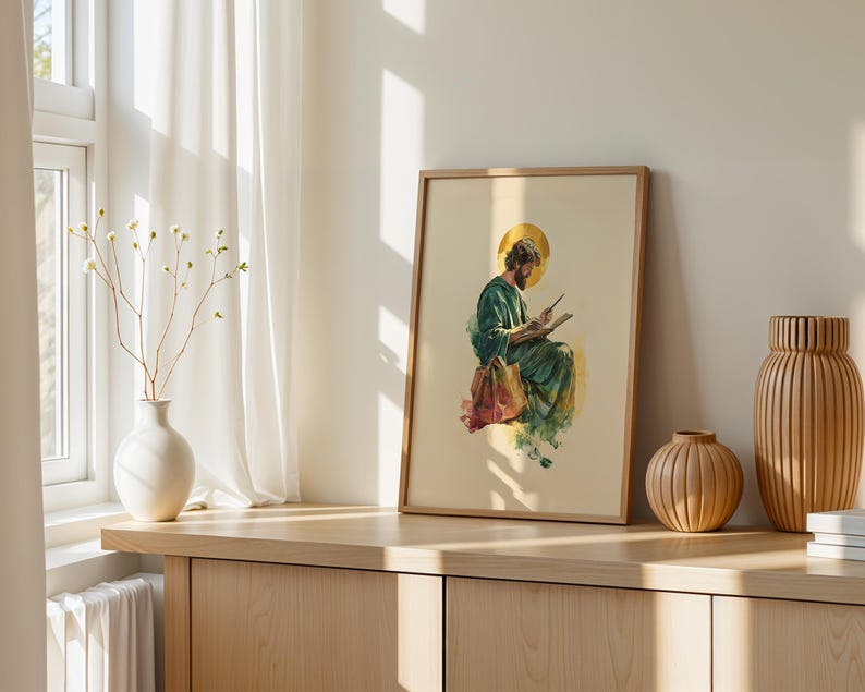 May include: A framed print featuring a watercolor illustration of a bearded figure in a green robe with a gold halo, sitting and writing in a book. The artwork is displayed in a light wood frame, positioned on a wooden surface next to vases.