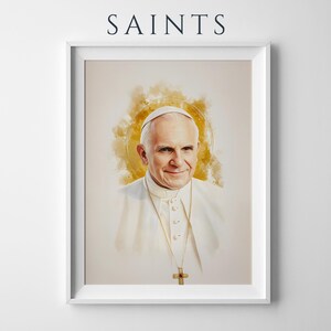 May include: A framed portrait print featuring the word SAINTS at the top. The artwork depicts a watercolour-style illustration of Pope John Paul II wearing a white cassock and a gold cross necklace, set against a soft, golden circular halo.