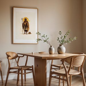 Op de afbeelding: Een ingelijste kunstprint met een figuur met een staf en een halo, boven een houten eettafel met twee lichtgekleurde keramische vazen met groene takken, vergezeld door drie houten stoelen met gevlochten zittingen.