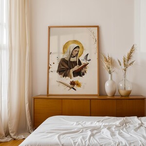 May include: A framed wall art print featuring a figure in a brown habit and white head covering writing in a book. A golden halo surrounds the head, with a small bird nearby. Below, two flowers and a quill are depicted on a cream background.