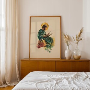 May include: A framed wall art print featuring a watercolor illustration of a bearded figure in a green robe with a golden halo, writing in a book. The figure sits with a brown bag nearby, rendered in soft green, yellow, and brown tones on a cream background.
