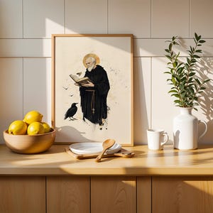 Op de afbeelding: Een ingelijste kunstdruk met een bebaarde monnik in een zwart habijt met een gouden aureool, die een boek leest met een raaf in de buurt. De print staat op een houten aanrecht naast een kom met gele citroenen, een wit bord, een houten lepel en een witte vaas.