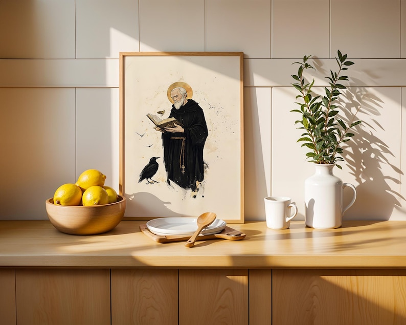 Op de afbeelding: Een ingelijste kunstdruk met een bebaarde monnik in een zwart habijt met een gouden aureool, die een boek leest met een raaf in de buurt. De print staat op een houten aanrecht naast een kom met gele citroenen, een wit bord, een houten lepel en een witte vaas.