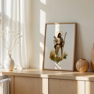 May include: A framed watercolour-style print depicting a figure with long hair and a beard, wearing robes and holding a small white lamb in one arm while carrying a wooden staff in the other hand, set against a soft, abstract golden halo background.