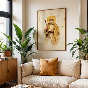 May include: A framed wall art print featuring a watercolor-style illustration of a bearded figure with a golden halo, wearing tan robes and holding a wooden staff. The artwork is displayed on a light-colored wall above a beige sofa with a mustard pillow.