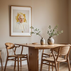 May include: A framed portrait of Pope John Paul II featuring a watercolour-style yellow background, displayed above a wooden dining table with two ceramic vases containing green branches, surrounded by four wooden chairs with woven seats.