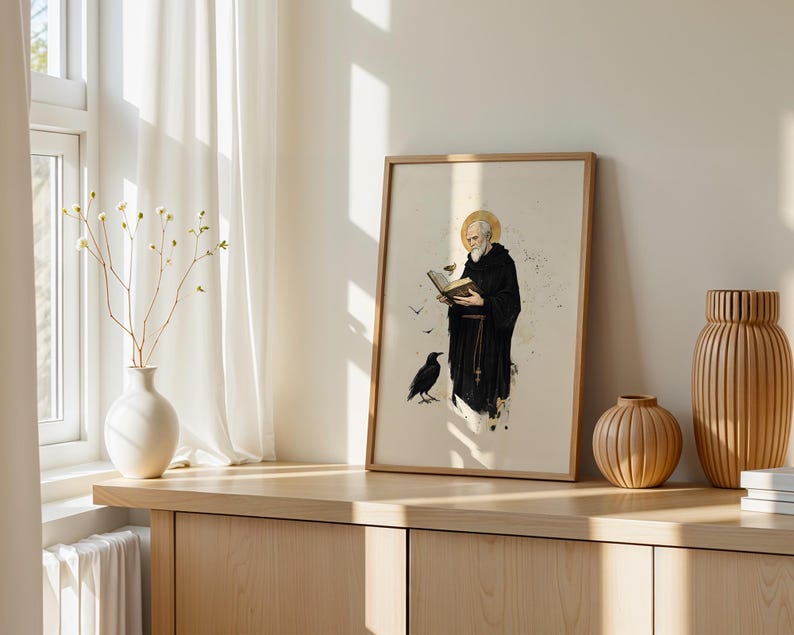 Op de afbeelding: Een ingelijste wandkunstprint met een illustratie van een bebaarde monnik in een zwart habijt die een open boek vasthoudt, met een gouden halo boven zijn hoofd en een zwarte raaf in de buurt, op een lichtcr&egrave;me achtergrond met inktspatten.