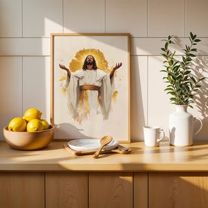 May include: A framed wall art print featuring a figure with long hair and a beard, wearing a white robe with a gold sash, arms raised, surrounded by a golden halo effect. The artwork is displayed in a light wood frame on a kitchen counter.