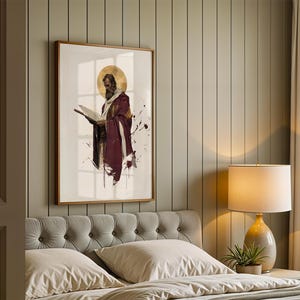 Op de afbeelding: Een ingelijste wandkunstprint met een bebaarde figuur in een dieprode mantel die een open boek vasthoudt, geaccentueerd door een gouden cirkelvormige halo en donkere inktachtige spetters op een witte achtergrond in een slaapkamer.