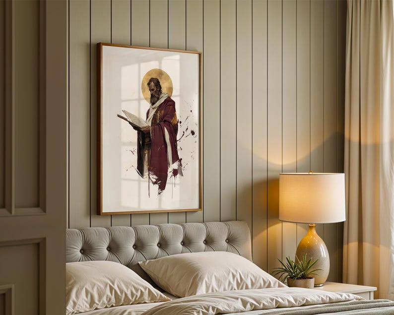 Op de afbeelding: Een ingelijste wandkunstprint met een bebaarde figuur in een dieprode mantel die een open boek vasthoudt, geaccentueerd door een gouden cirkelvormige halo en donkere inktachtige spetters op een witte achtergrond in een slaapkamer.