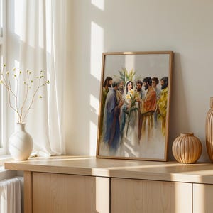 May include: A framed watercolor print depicting a group of figures in robes surrounding a central woman holding yellow lilies. The artwork features soft, blended colors and is displayed in a light wood frame on a wooden surface.