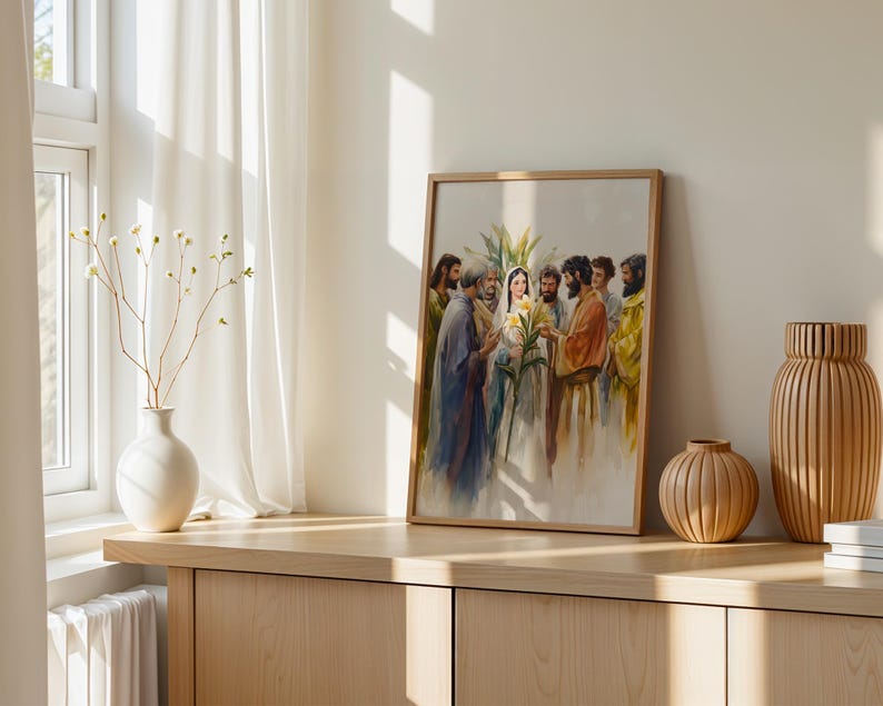 May include: A framed watercolor print depicting a group of figures in robes surrounding a central woman holding yellow lilies. The artwork features soft, blended colors and is displayed in a light wood frame on a wooden surface.