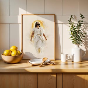 May include: A framed wall art print depicting a figure in white robes with a golden halo, positioned above a wooden counter with a bowl of yellow lemons, a white plate, a wooden spoon, a white mug, and a white vase containing green foliage.