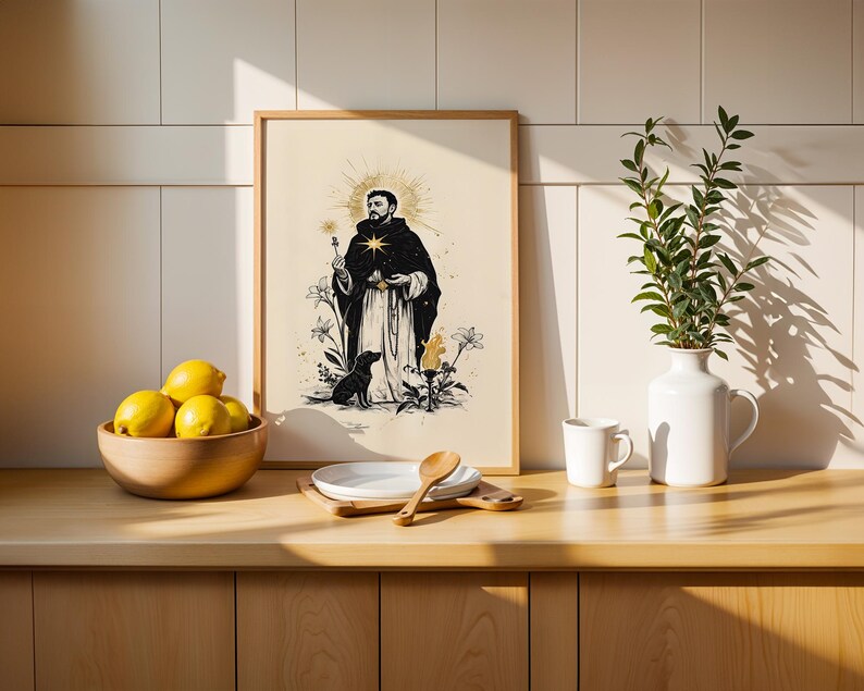 May include: A framed art print featuring a black and white illustration of a robed figure with a starburst halo, a dog, and floral elements. The print is displayed on a wooden counter next to a bowl of yellow lemons, a white plate, and a vase.