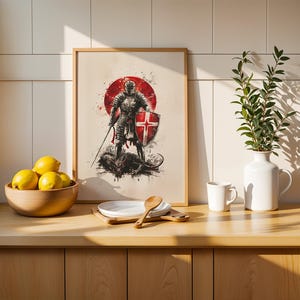May include: A framed wall art print featuring a knight in full armour holding a sword and a shield with a red cross, standing over a dark creature against a red circular background. The artwork is displayed on a wooden counter next to a bowl of lemons.