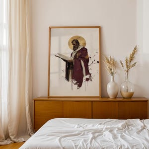 May include: A framed vertical wall art print featuring a bearded figure in a deep purple robe with a gold halo, reading an open book. The artwork uses a watercolour style with ink splatters on a white background, displayed in a light wood frame.