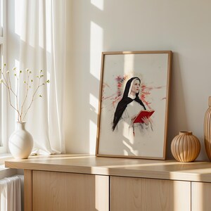 May include: A framed wall art print featuring a portrait of a nun wearing a black and white habit, holding an open red book. Behind the figure is a stylized red and orange watercolor-style burst. The artwork is presented in a light wood frame.