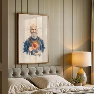 May include: A framed wall art print featuring a watercolor-style portrait of a bearded individual wearing blue vestments with a glowing, fiery heart emblem on the chest and a golden halo, presented in a simple wooden frame on a paneled wall.