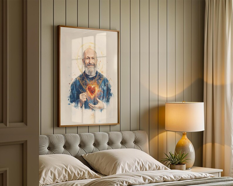May include: A framed wall art print featuring a watercolor-style portrait of a bearded individual wearing blue vestments with a glowing, fiery heart emblem on the chest and a golden halo, presented in a simple wooden frame on a paneled wall.