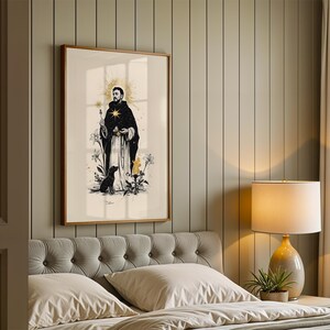 Op de afbeelding: Een ingelijste verticale wandkunstprint met een zwart-witillustratie van een figuur in een gewaad met een gouden sterrenhalo, die een klein voorwerp vasthoudt, vergezeld door een zwarte hond, lelies en een kaars, alles tegen een lichte achtergrond.