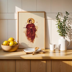 May include: A framed art print featuring a figure in a deep red robe with a golden halo, displayed on a wooden counter. The print is surrounded by a wooden bowl of yellow lemons, a white plate with a wooden spoon, a white mug, and a white vase.