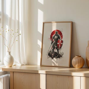 May include: A framed wall art print featuring a knight in full armour holding a sword and a shield with a red cross, standing over a dark creature. The illustration uses a red circular background with ink splatter effects on a light background.