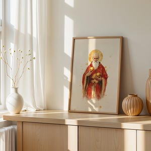May include: A framed vertical print featuring a watercolor illustration of a bearded figure in red robes with a golden halo, holding two golden spheres. The artwork is displayed in a light wood frame, positioned on a wooden surface with vases.