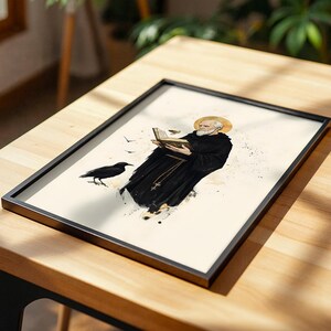Op de afbeelding: Een ingelijste print met een illustratie van een bebaard figuur in een zwart gewaad die een open boek vasthoudt, met een gouden halo boven het hoofd en een zwarte raaf ernaast, op een cr&egrave;mekleurige achtergrond met inktspatten.