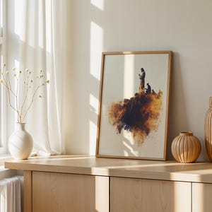 May include: A framed wall art print featuring a watercolour-style illustration of a figure standing on a rocky cliff edge, rendered in shades of brown, gold, and dark blue, set within a light-toned wooden frame on a flat surface.