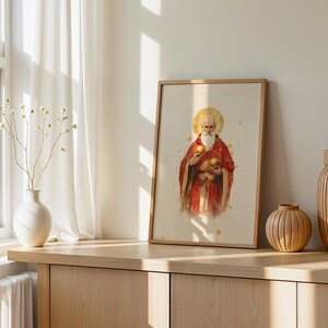 May include: A framed wall art print featuring a watercolour-style illustration of a bearded figure in red robes with a golden halo, holding two golden spheres. The artwork is displayed in a light-coloured wooden frame on a wooden surface.