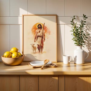 Peut inclure: Une impression encadr&eacute;e de style aquarelle repr&eacute;sentant une silhouette barbue en robe marchant avec un petit agneau sur fond de soleil orange circulaire. L'&oelig;uvre est pos&eacute;e sur un comptoir en bois &agrave; c&ocirc;t&eacute; d'un bol de citrons et d'un vase blanc.