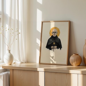 May include: A framed art print featuring a digital illustration of a bald person wearing glasses and a dark hooded robe. The figure has a golden circular halo behind their head and holds a rosary with a cross pendant against a cream background.