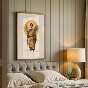 Op de afbeelding: Een ingelijste kunstprint met een figuur in gouden gewaden voor een grote cirkelvormige halo. De figuur houdt een hoge staf en een palmblad vast. Het kunstwerk is ingelijst in een houten lijst aan een verticaal beklede wand boven een gecapitonneerd hoofdbord.