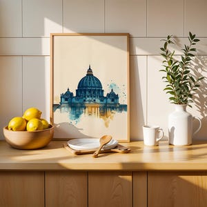 May include: A framed watercolour-style print depicting the dome of St. Peter's Basilica in shades of blue and gold. The artwork is displayed on a wooden counter next to a wooden bowl filled with yellow lemons, a white plate, a spoon, and a white vase.