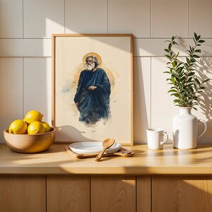 May include: A framed art print featuring a watercolor illustration of a bearded figure in a dark blue robe with a golden halo. The print is displayed alongside a wooden bowl of yellow lemons, a white plate with a wooden spoon, and a white vase.