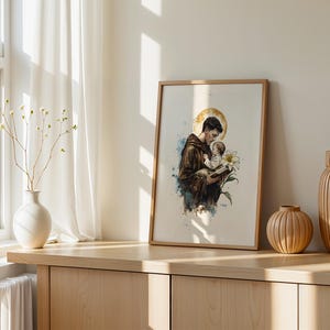 Puede incluir: Una l&aacute;mina enmarcada de estilo acuarela que representa a una figura con h&aacute;bito marr&oacute;n sosteniendo a un ni&ntilde;o peque&ntilde;o, con un halo dorado, un lirio blanco y salpicaduras de pintura azul. La obra se presenta en un marco de madera clara.