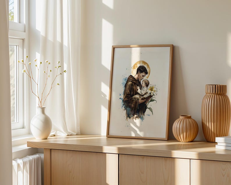 Puede incluir: Una l&aacute;mina enmarcada de estilo acuarela que representa a una figura con h&aacute;bito marr&oacute;n sosteniendo a un ni&ntilde;o peque&ntilde;o, con un halo dorado, un lirio blanco y salpicaduras de pintura azul. La obra se presenta en un marco de madera clara.