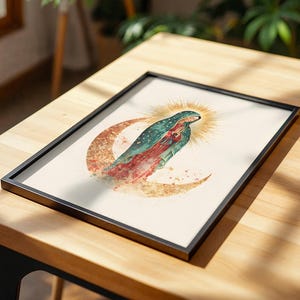 May include: A framed print featuring a watercolor illustration of the Virgin of Guadalupe standing on a crescent moon. The figure wears a teal mantle with gold stars over a red dress, surrounded by a golden sunburst halo on a cream background.