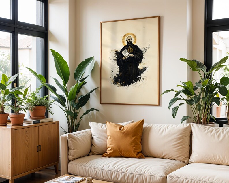 May include: A framed wall art print featuring a stylised illustration of a monk in a black habit with a gold halo, holding a white document. The artwork uses a cream background with black ink-like splatters and gold accents around the figure.