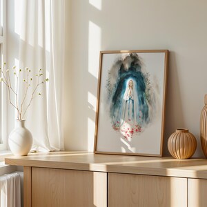 Puede incluir: Una l&aacute;mina de acuarela enmarcada que representa una figura con t&uacute;nica blanca y azul con las manos juntas en oraci&oacute;n, rodeada por un fondo abstracto azul oscuro y gris con detalles florales rojos en la base, en un marco de madera clara.