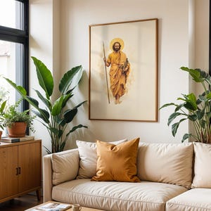 Op de afbeelding: Een ingelijste kunstdruk met een bebaarde figuur in een gouden gewaad die een lange staf en een boekrol vasthoudt, met een cirkelvormige halo achter het hoofd. Het kunstwerk hangt aan een lichte muur boven een beige bank met een mosterdkleurig kussen.