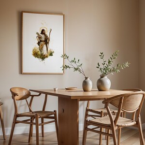 May include: A framed watercolour-style wall art print depicting a figure holding a lamb and a staff, displayed above a wooden dining table with two ceramic vases, three wooden chairs, and a small wooden stool with a stack of plates.
